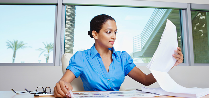Woman with a blue shirt reading a long printout with her glasses resting beside her hand on a desk