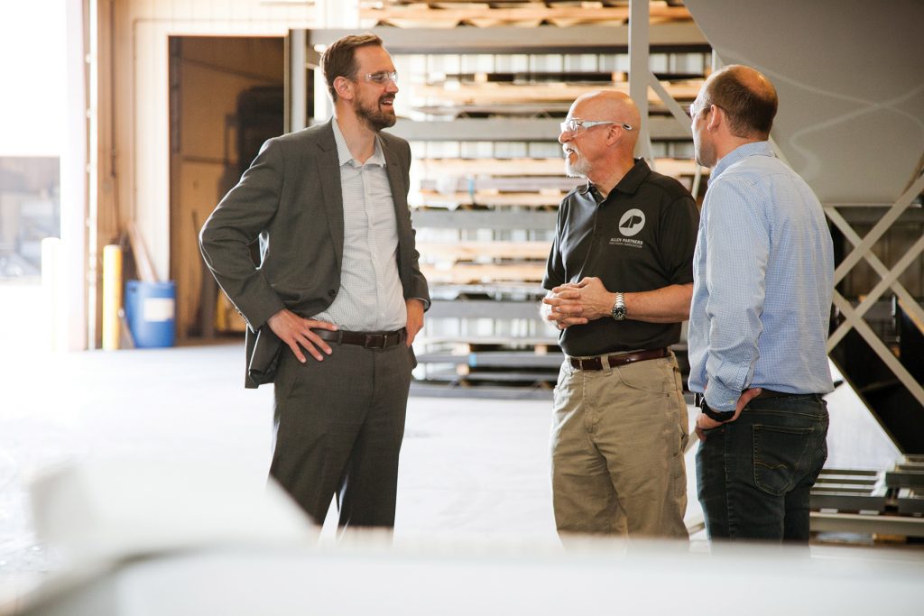 Three men wearing safety glasses in a warehouse conversing with each other
