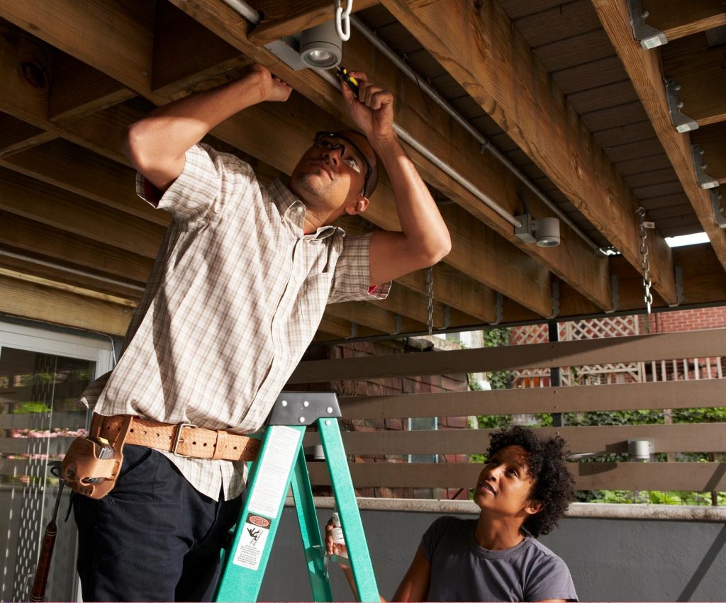 Man on a ladder installing track lighting under a deck with a woman