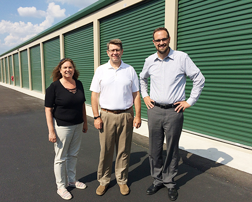 Two men and a woman standing outside of storage unit facility with green doors on a sunny day