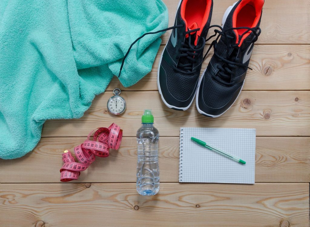 A pair of running shoes, bottle of water, measuring tape, stopwatch, pen and paper, and a green towel