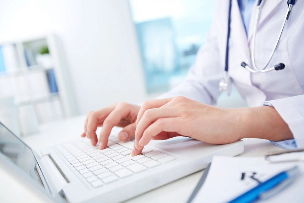 A close-up of a doctor typing on a white laptop in a clinical setting