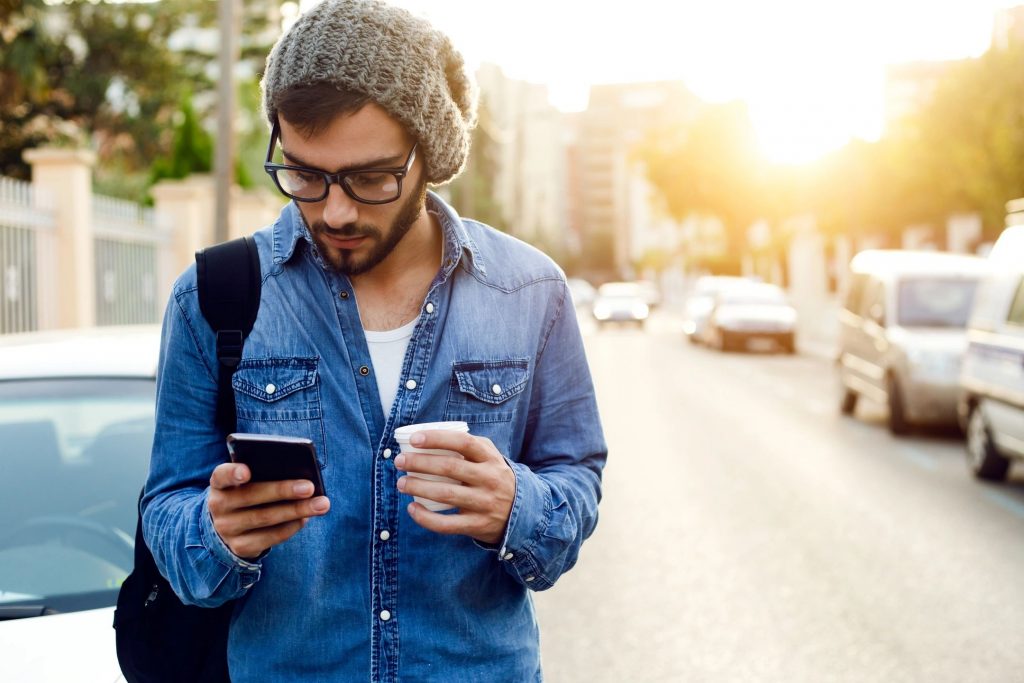 A young man with a denim shirt and snow hat with a coffee and a cellphone standing in the street