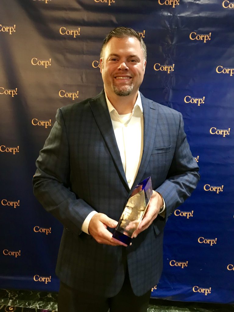 A man smiling holding an award in front of a step-and-repeat at a Corp! event