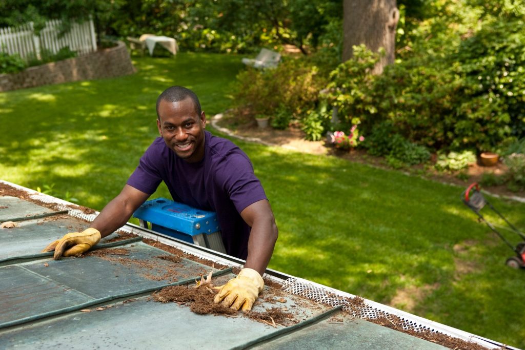A man on a ladder cleaning leaves and pine needles off a metal roof and a gutter