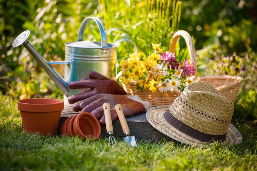 A pile of gardening equipment: flower pots, watering can, gardening glove, small spades, hat, and a basket with freshly picked flowers