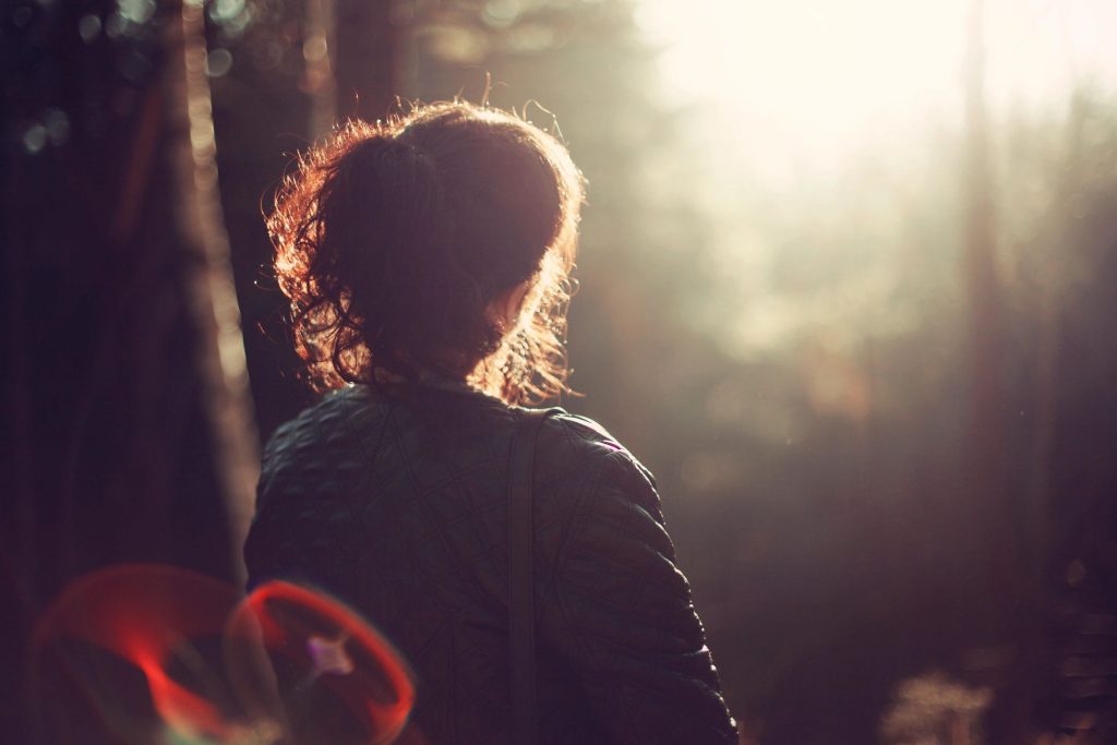 A woman with brown hair from behind in a wooded area with the sun filtering through