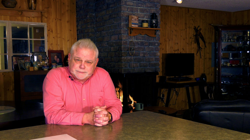A man with a pink shirt sitting in a living room with a fire going in the background