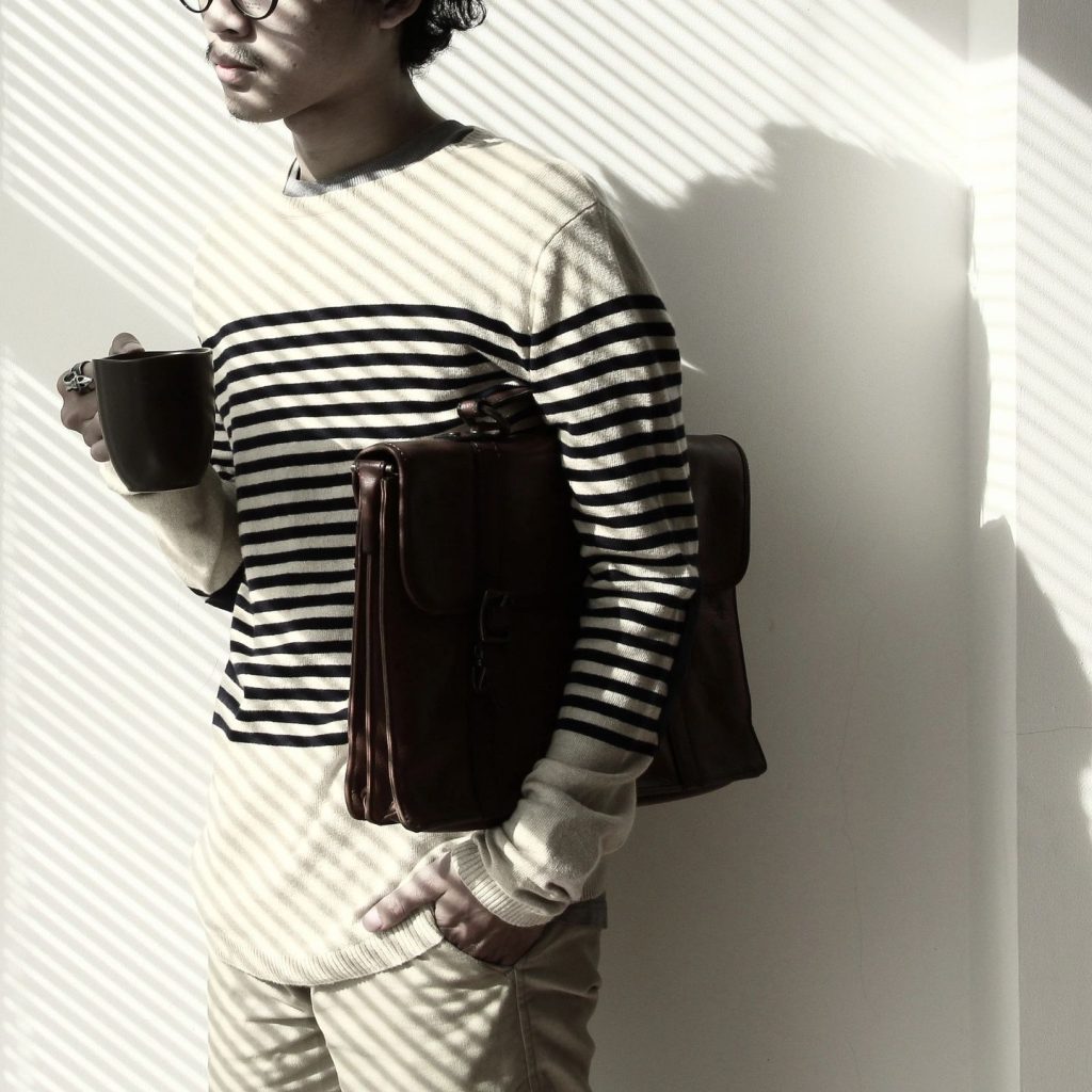 A young man standing against a white wall holding a coffee cup with a briefcase under his arm wearing a striped sweater