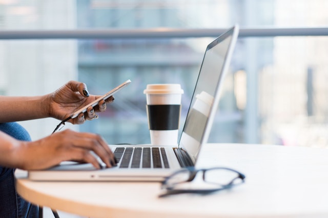 A close-up of a person on their laptop and cellphone with a coffee and a pair of glasses on the table