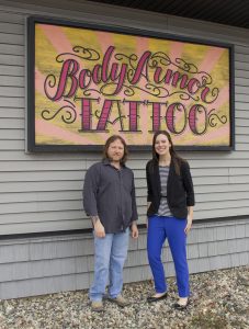 A man and a woman standing in front of a Body Armor Tattoo sign