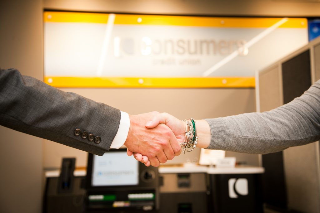 A man and a woman shaking hands in front of a Consumers Credit Union sign on an office wall