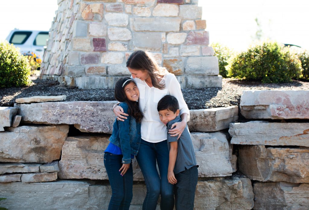 A woman embracing a boy and a girl in front of large stones and a brick structure