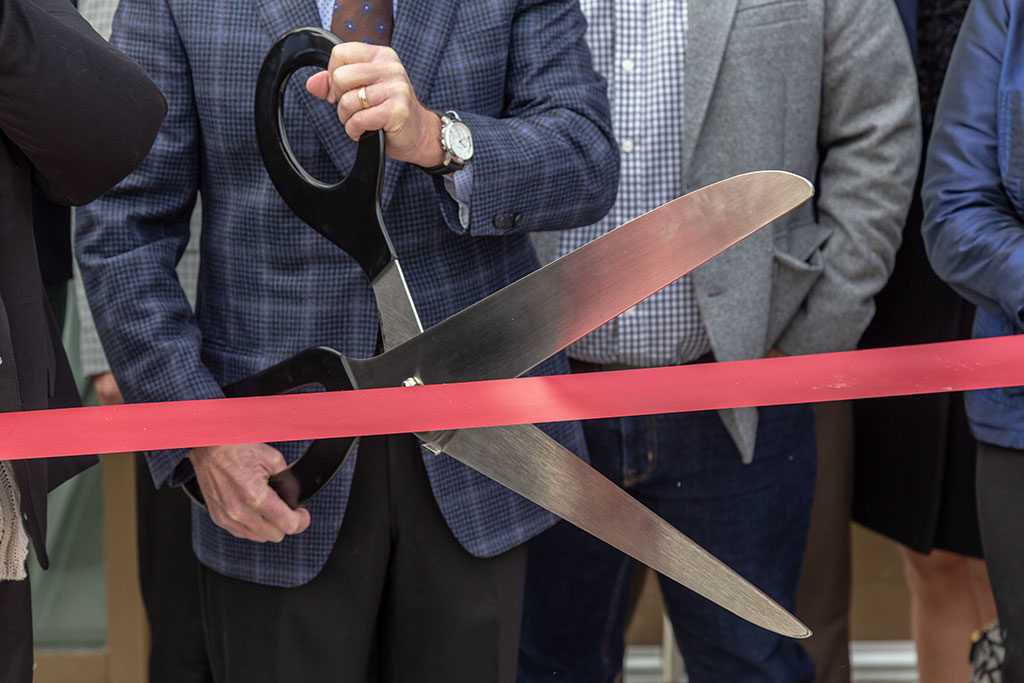 A man with a blue plaid sport jacket holding an oversized pair of scissors cutting red ribbon.
