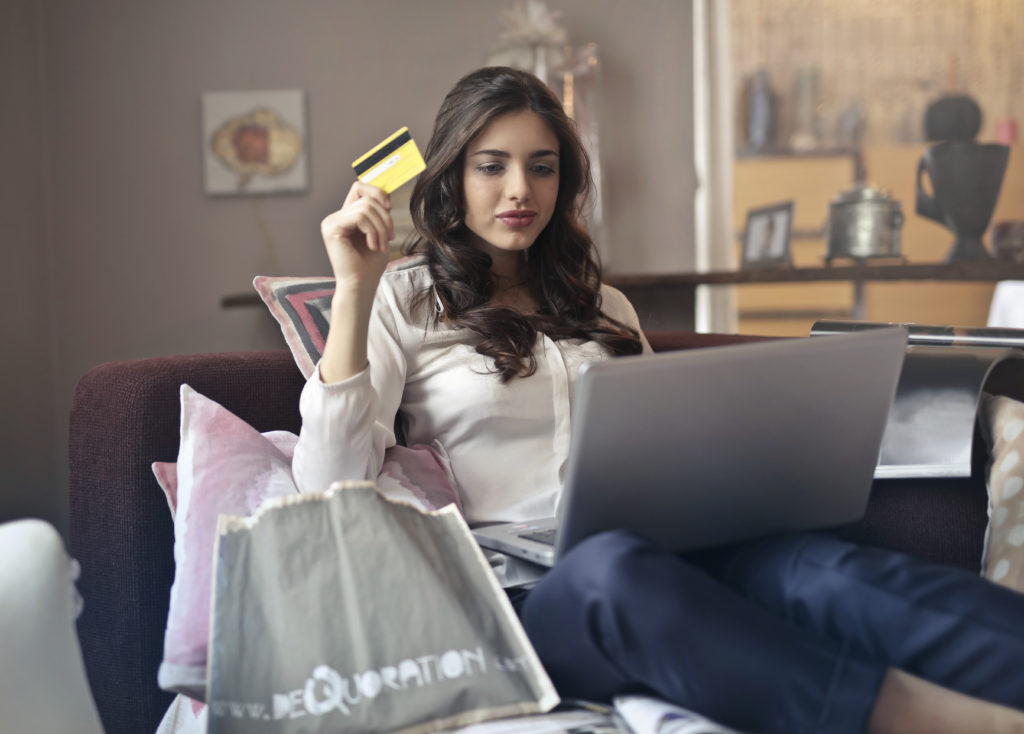 A woman on her couch holding a yellow credit card buying something on her laptop