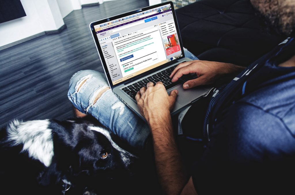 A man checking his email on a laptop while sitting on his couch next to his dog