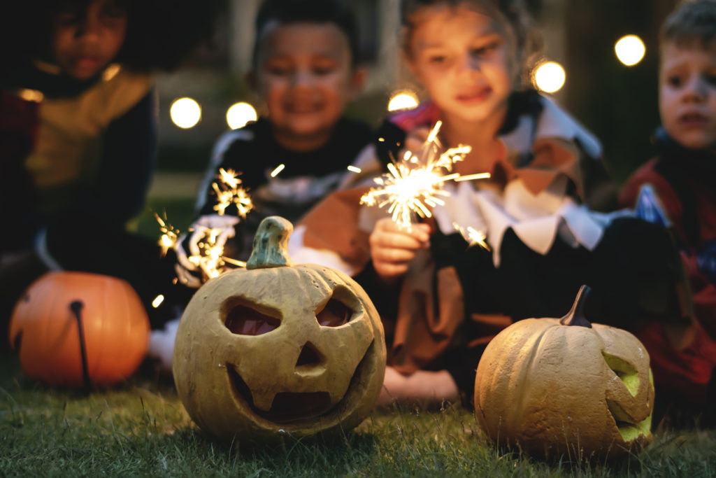 Three children in costumes holding sparklers near ceramic jack-o'-lanterns.