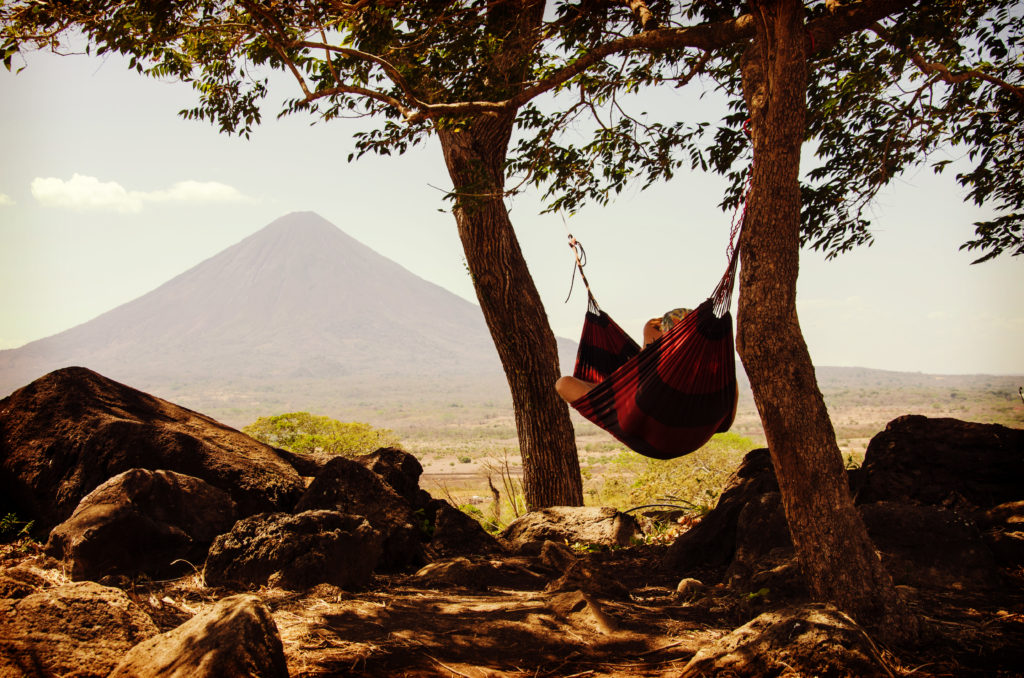 A person lounging in a red hammock under some trees in front of a mountain.