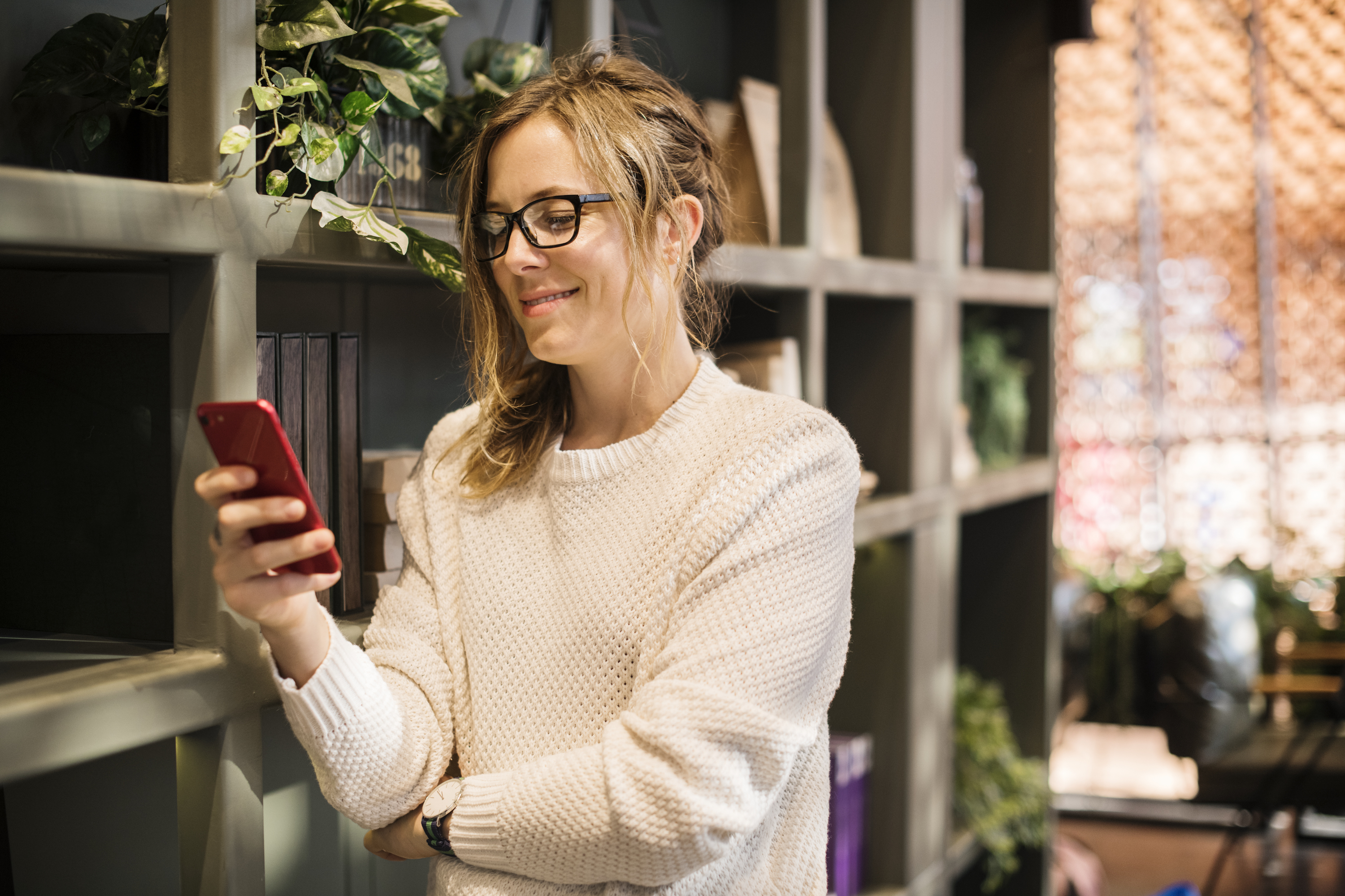 A woman wearing glasses and a white knit sweater holding a red mobile phone in front of bookshelves.