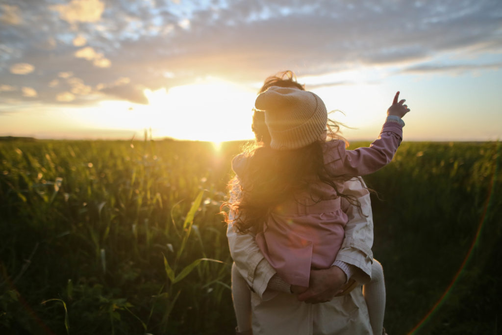 A mother holding her daughter wearing a pink overcoat standing in tall grass at sunrise.