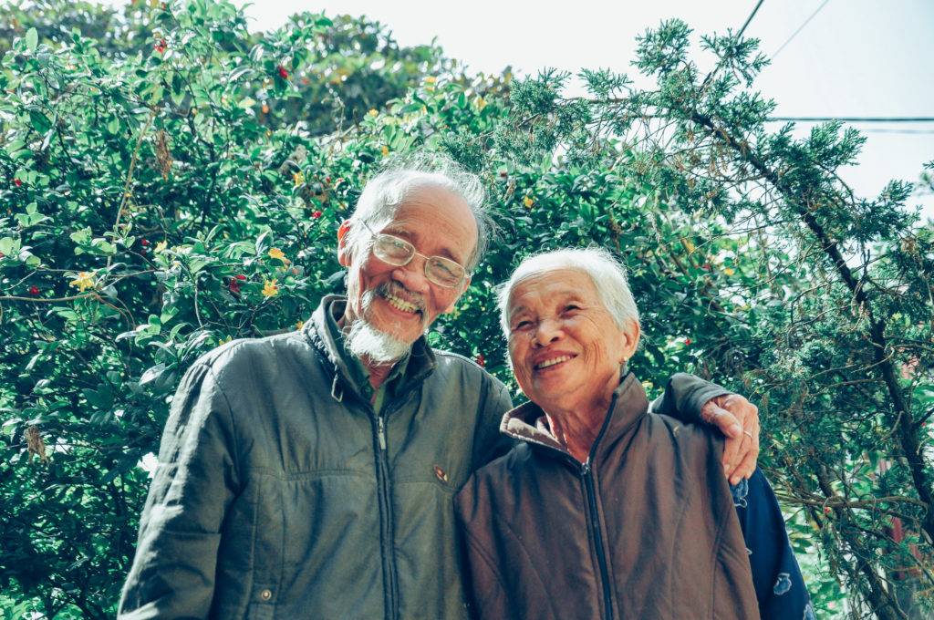 An older couple arm in arm standing under a tree blossoming red and yellow flowers.