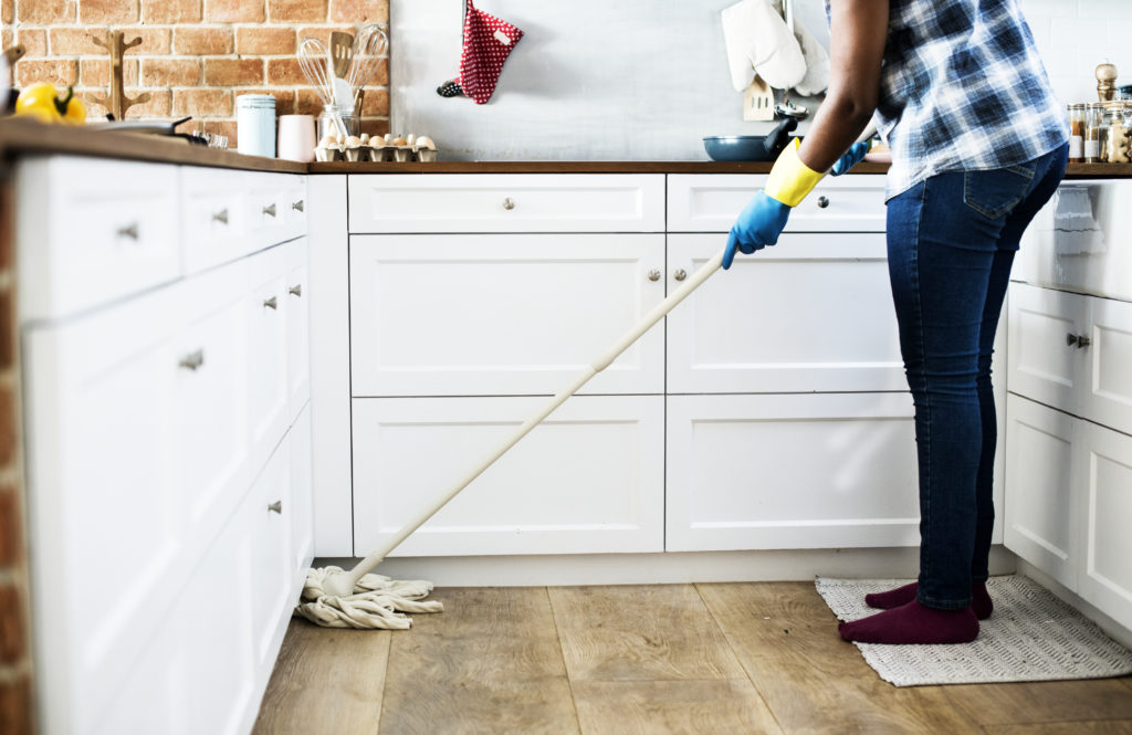 Someone mopping wood floor in a kitchen with white cabinets and brick walls.