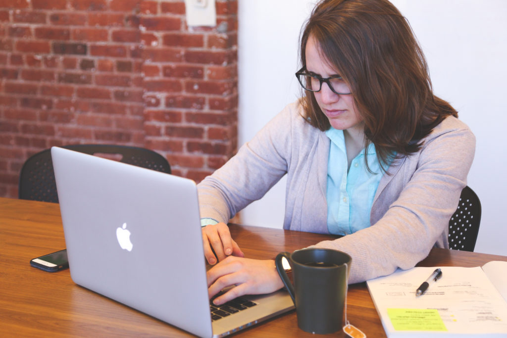 A woman wearing glasses drinking tea and using a Macbook sitting at a table.
