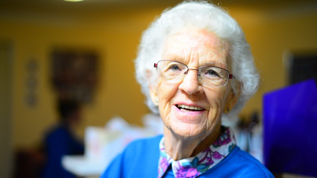 An elderly lady with a floral shirt and a bright blue sweater smiling.