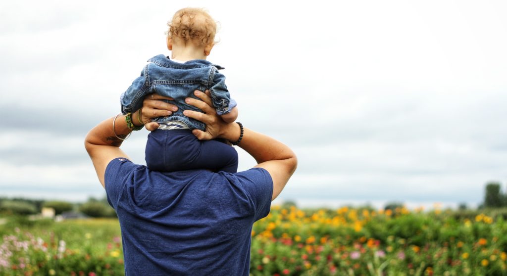 A father holding a baby on his shoulders in a field of flowers on a cloudy day.
