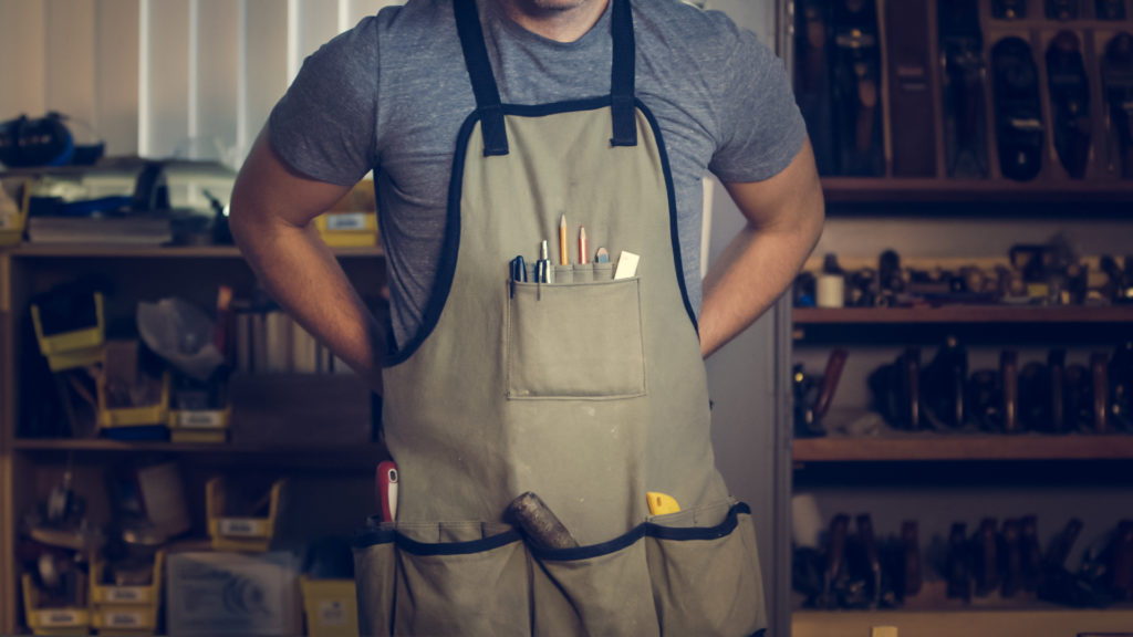 A man wearing a work apron in a workshop with a wall of tools and supplies on shelves.