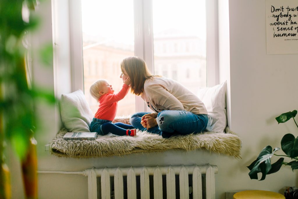 A mother sitting with a toddler on a furry blanket in a bay window playing.