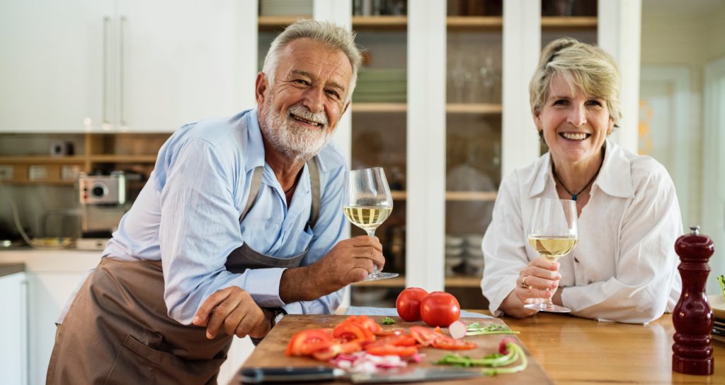Married couple holding glasses of white wine over a island countertop with a cutting board covered with chopped vegetables.