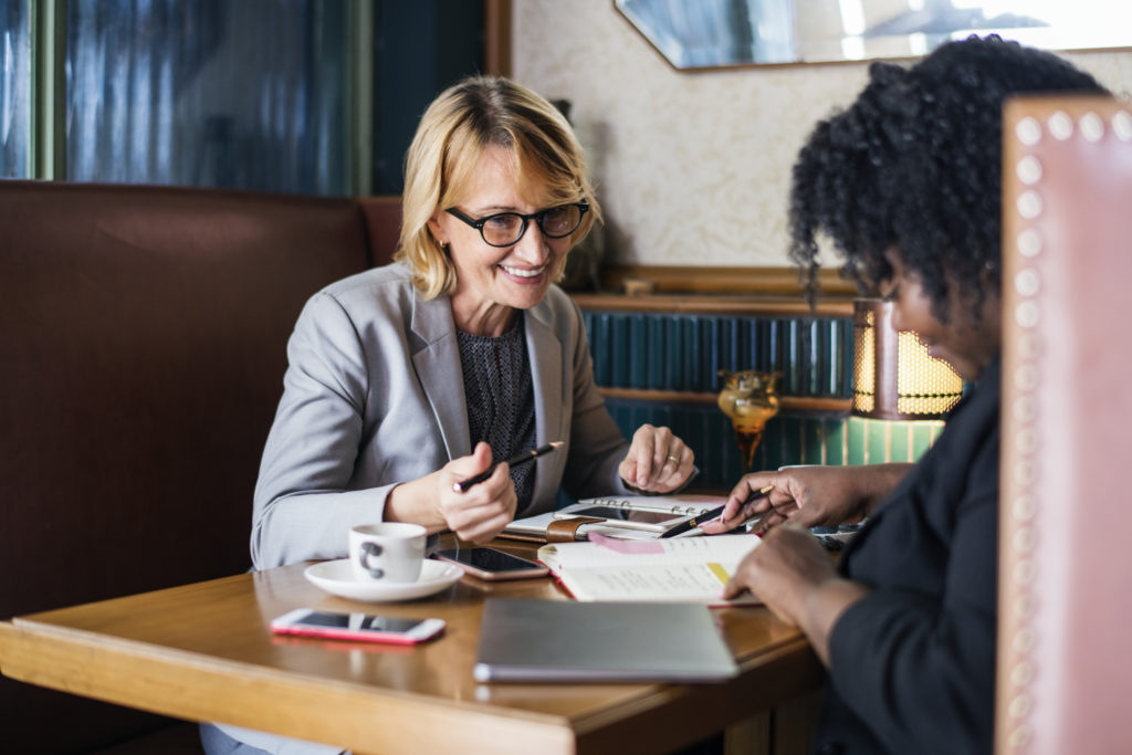 Two women sitting a booth referring to their agendas while enjoying coffee.