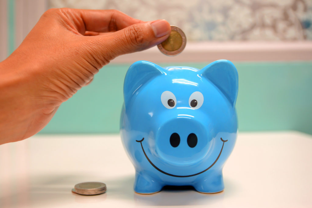 A hand placing British currency into a blue piggy bank.