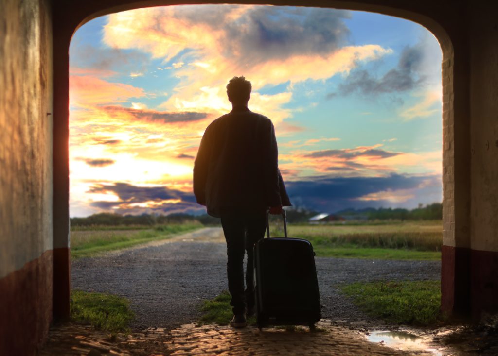 A person with a rolling suitcase walking out of a garage looking out over a rural area at sunrise.