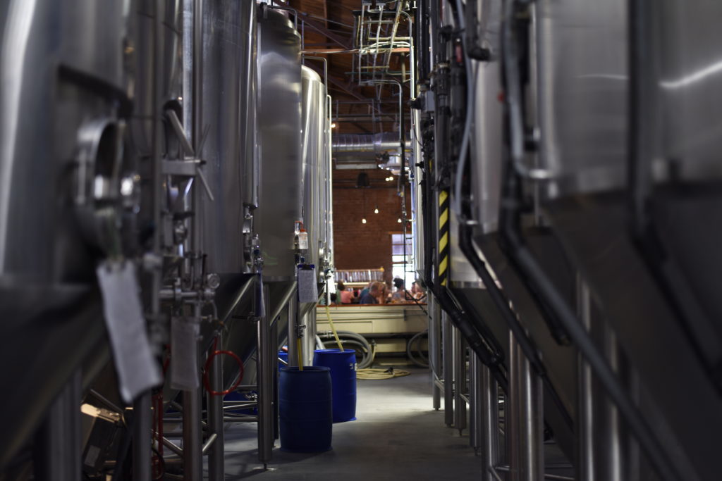 Rows of large metal vats at a brewery with customers seated in the background in the bar/dining area.