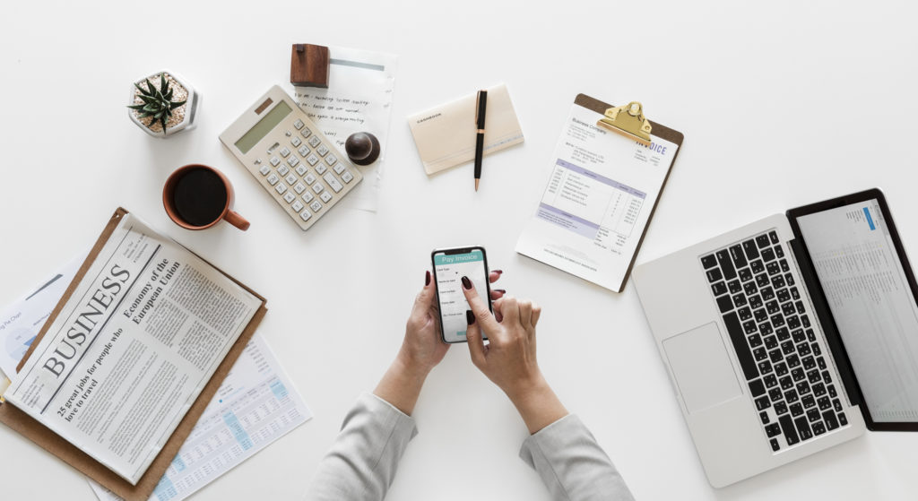 A person holding a cellphone over a white table with a clipboard, pen, calculator, coffee, succulent, and laptop.