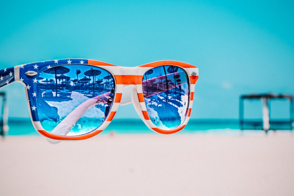 A pair of sunglasses with an American Flag design on the frame on a beach.