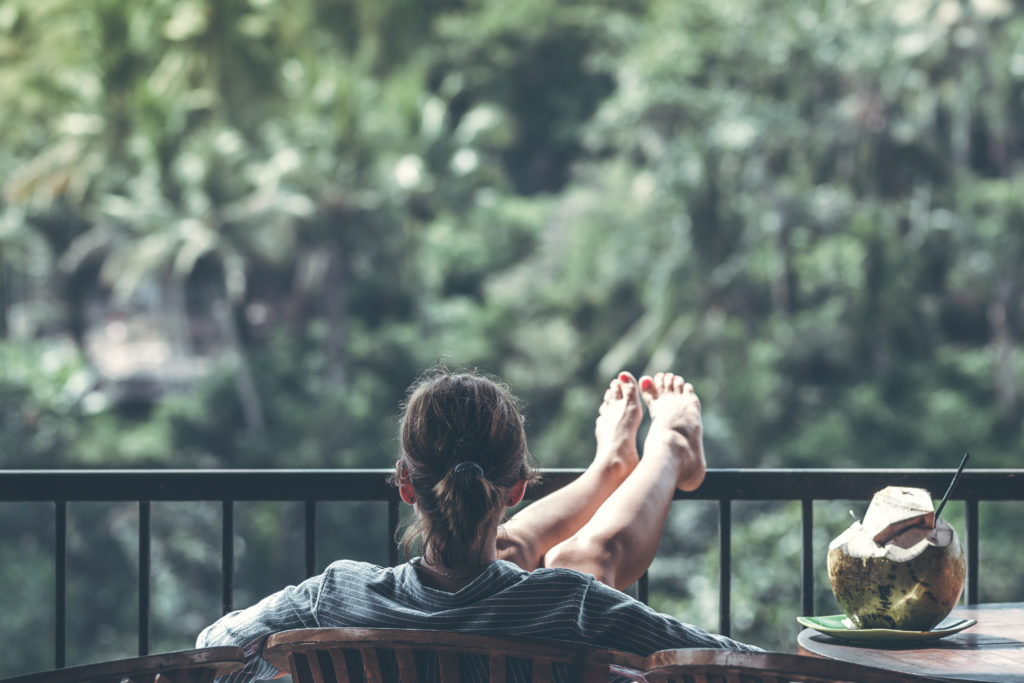 A person reclining in a wooden chair on a balcony with a coconut drink on a table next to them.