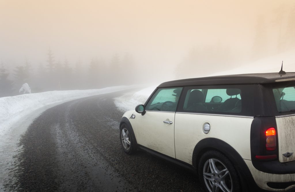 A white Mini Cooper driving down a snowy, winding road on a foggy winter day.
