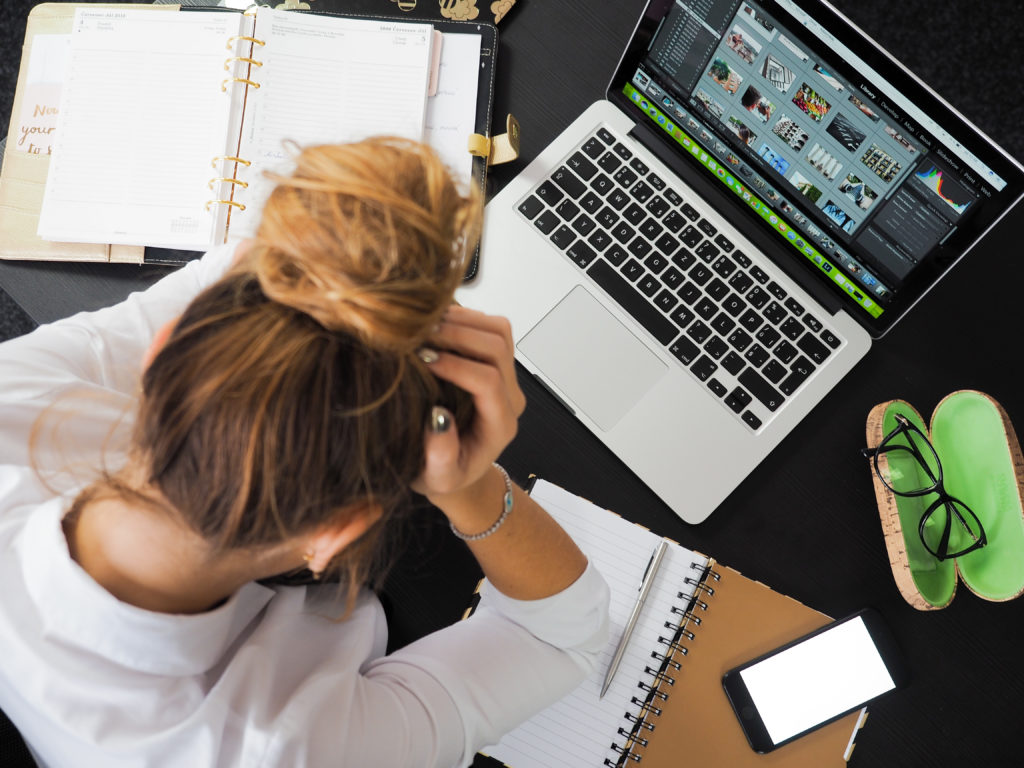 A woman with her head in her hands over a laptop, planner, and notebook looking stressed out.