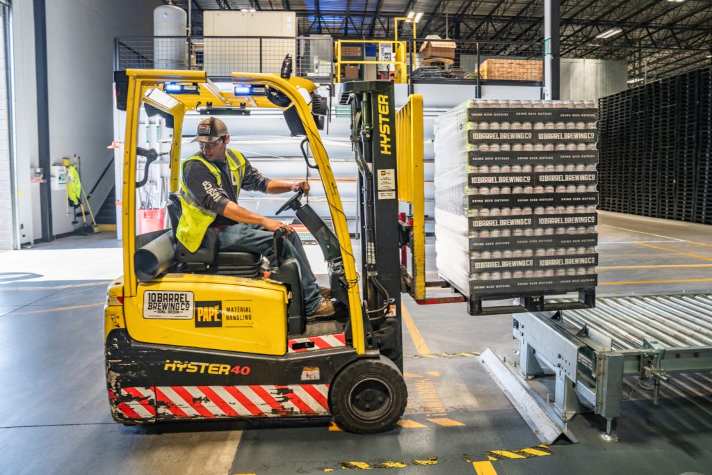 A forklift operator in a warehouse carrying a pallet of beer cans.