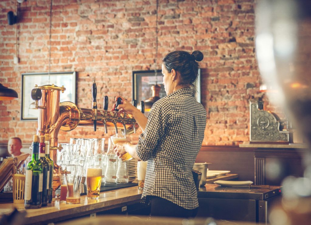 A bartender wearing a black and white gingham shirt pouring a draft beer at a bar.