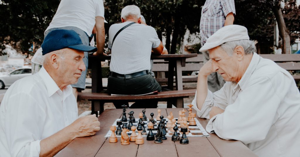 Two elderly gentlemen playing chess on a picnic table in a park.
