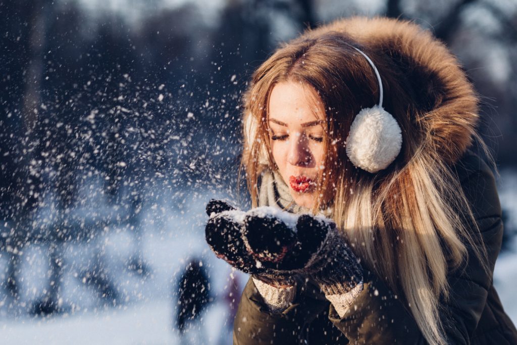 A young woman wearing a parka, earmuffs, and mittens blowing on a pile of snow cupped in her hands.