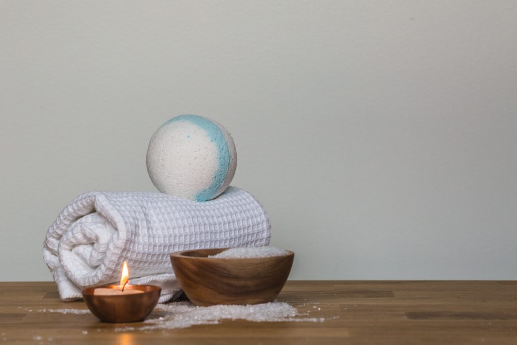 A bath bomb resting on a rolled towel in by a wooden bowl of bath salts next to a candle.