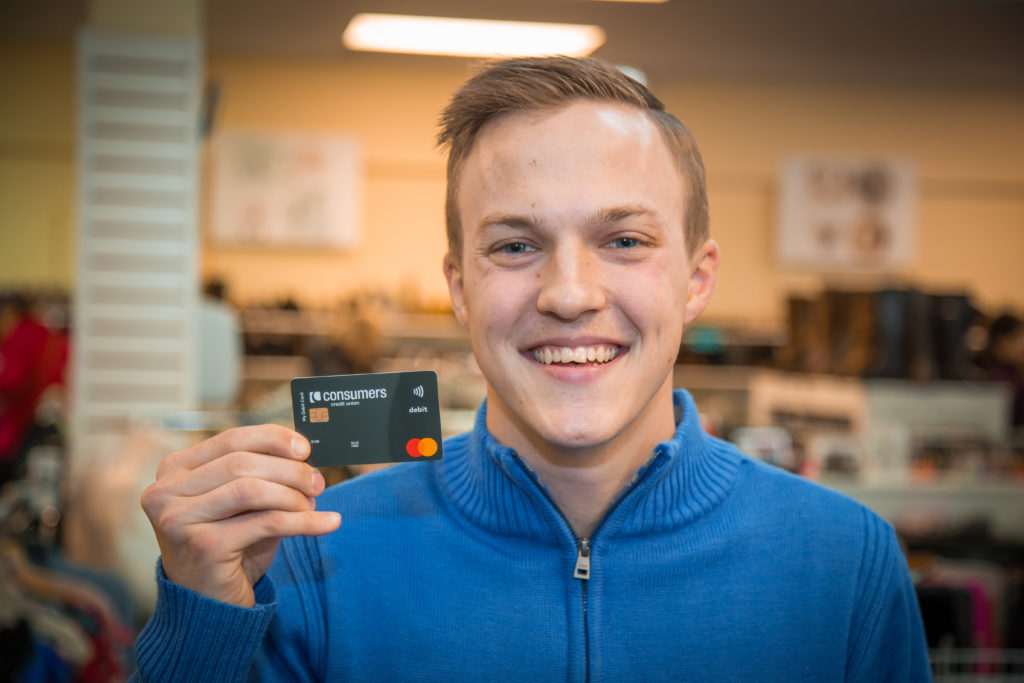 A blonde young man wearing a blue sweater holding a black Consumers Credit Union debit card.