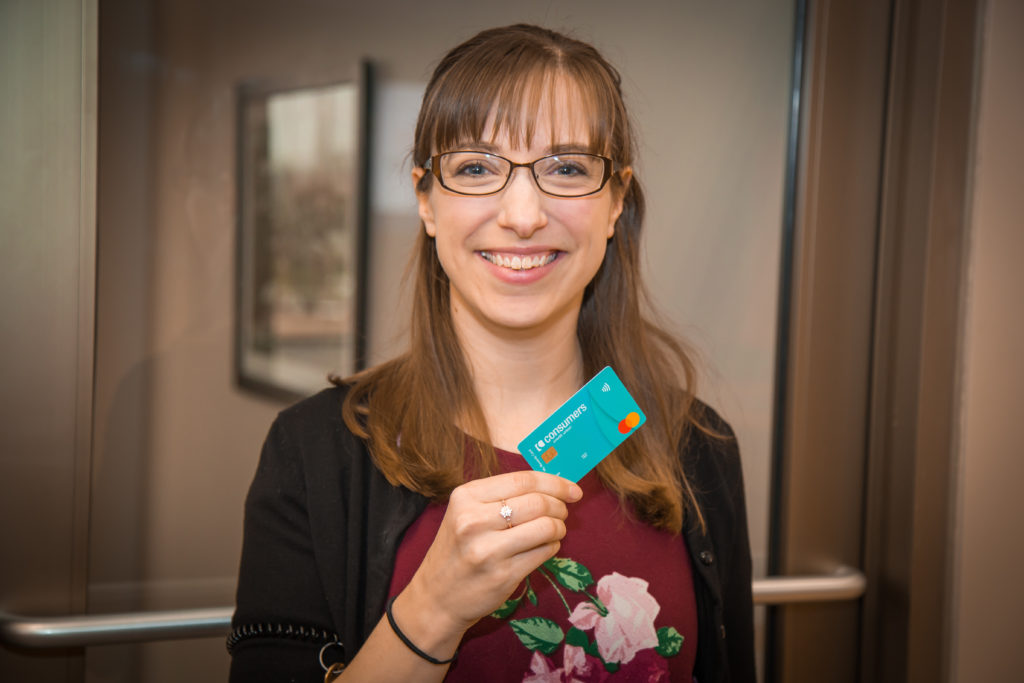 A woman with glasses and a cardigan holding a blue Consumers Credit Union Rewards card.