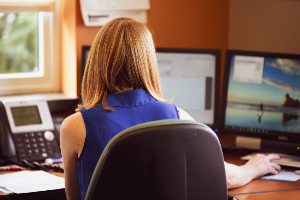 A woman wearing a royal blue sleeveless shirt at a desk working from behind.