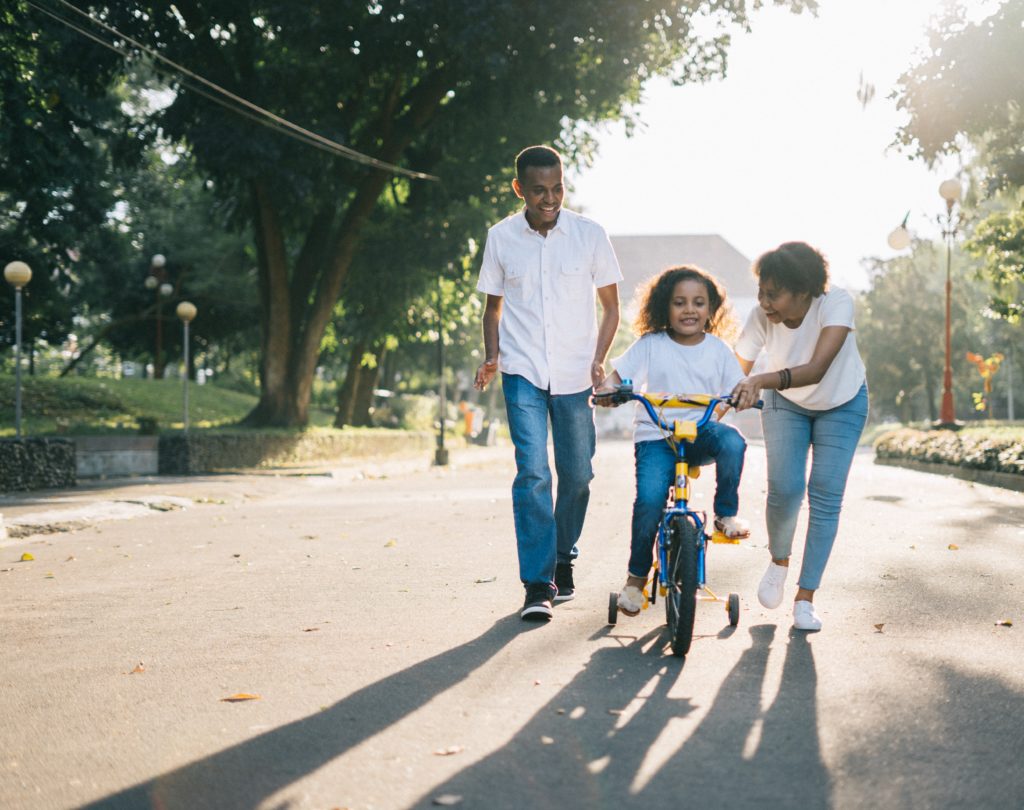 Man standing beside his wife teaching their child how to ride a bike.